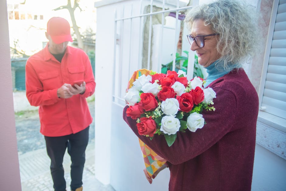 A woman with curly gray hair and glasses, wearing a maroon sweater and blue turtleneck, is holding a vibrant floral bouquet featuring red and white roses with green foliage, wrapped in colorful patter