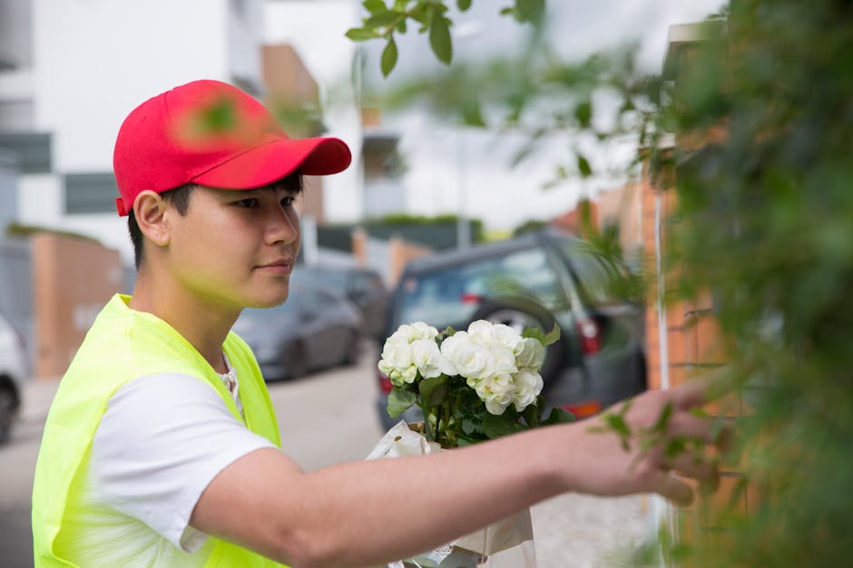 A young delivery person wearing a red cap and a high-visibility yellow vest is placing a floral gift, a bouquet of white hydrangeas, against a brick wall. The bouquet is wrapped in clear plastic with 
