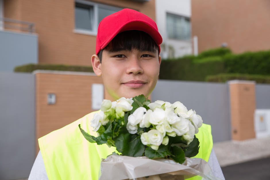 A young male delivery person wearing a red cap and a high-visibility yellow vest is standing outdoors in a residential area with modern brick and concrete houses and neatly trimmed greenery in the bac
