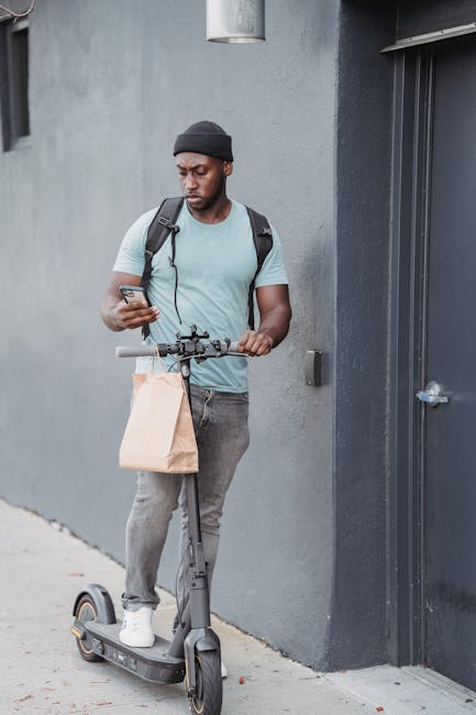 A young man with dark skin, wearing a black beanie, light blue t-shirt, and grey jeans, stands outdoors against a grey concrete wall. He holds a smartphone in his right hand and grips the handlebars o