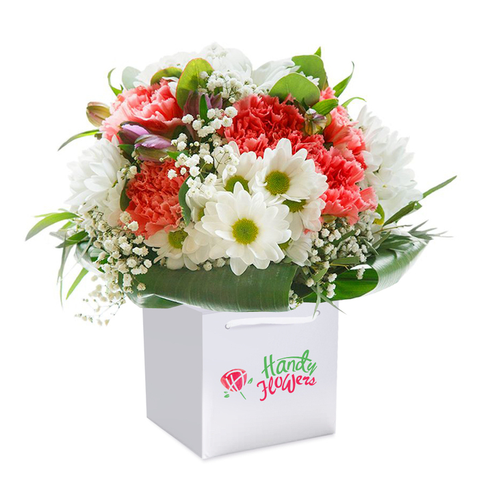 Brightening its pristine white surroundings, this bouquet is a celebration of vivid colour and texture inspired by the natural beauty of Chessington. The heart of the ensemble features sumptuous coral and peach carnations, their undulating petals inviting touch and admiration, clustered with pure white daisies whose glowing lime centers lend vitality. Wisps of baby's breath float delicately between the dominant blooms, imparting an airy elegance and softening the bouquet's overall silhouette. Discreetly woven in on one side, several unopened buds with purple-tinged tips provide an intriguing counterpoint, hinting at the promise of blooms yet to unfold. Lush, deep-green leaves frame the arrangement, their broad, lustrous surfaces creating both structure and visual harmony. Everything is presented in a crisp white square gift bag, the handles peeking out for effortless carrying-a nod to the convenience favoured by Chessington Florist clients. Photographed against a flawless white background, each colour and texture is rendered in sharp relief. From casual tokens of appreciation to momentous events like birthdays or anniversaries, this thoughtfully designed bouquet is ready to bring joy and a touch of local artistry to homes across Chessington and Malden Rushett.