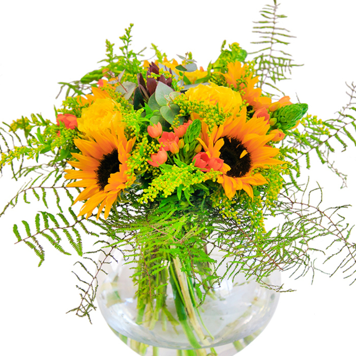 The initial impression of this Chessington Florist bouquet is pure, sunlit exuberance-an artful expression of classic garden style with a modern twist. Vivid sunflowers with rich, chocolate-brown hearts and golden petals come together with plush yellow garden roses, whose tightly furled petals spiral like a fairy-tale rose in full bloom. Scarlet Hypericum berries are woven through the arrangement, their glossy surfaces shining amidst layers of green-a playful visual note that contrasts perfectly with the softness of the petals. Cascading fern fronds add feathery movement and a wild, outdoorsy touch, echoing the breezy trails winding through nearby Chessington parks. Airy clouds of light green solidago and slender green filler spikes provide seasonal texture and dimension, while muted eucalyptus leaves soften the palette with their silvery blue-green warmth. The whole arrangement is anchored in a crystal-clear, round glass vase, its transparent body offering a window into the natural beauty and architectural structure of the bouquet. Light filters across the display, making each stem and leaf glow. A gentle scent of fresh roses and greenery completes the sensory picture, reminiscent of a stroll near Chessington's wooded paths. Designed to delight for birthdays, anniversaries, or everyday uplifting moments along Leatherhead Road, this bouquet is a true celebration of nature's vibrant palette and the skill of local florists.